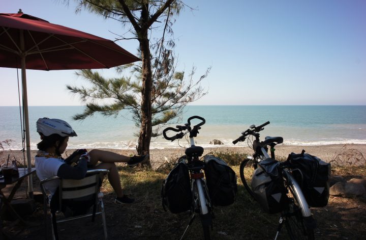 A cyclist resting under a beach umbrella with two bicycles.
